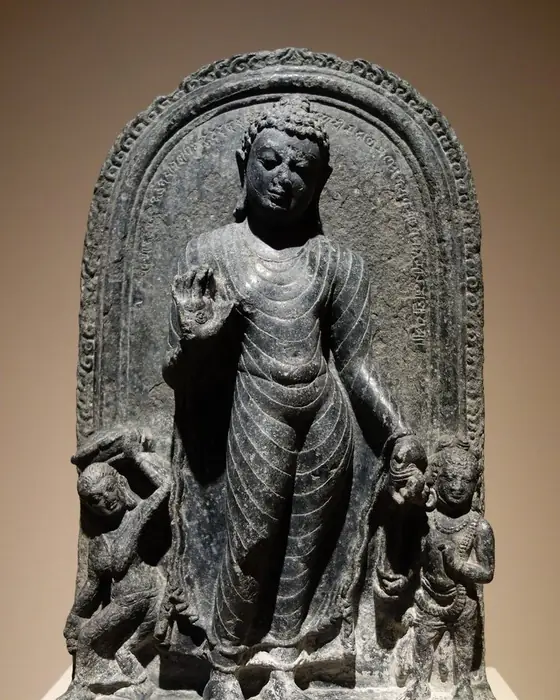 A dark basalt stele of the standing Buddha, with small figures at his feet.