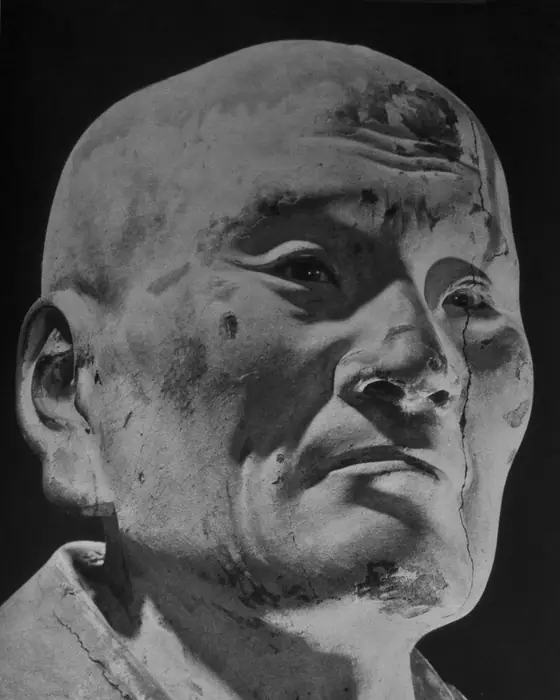 A black-and-white photograph of a wooden statue of an old monk with a lined, attentive face.