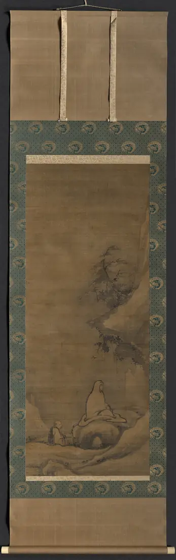 A tall hanging scroll with a small seated figure before a dark cliff.