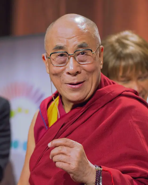 A colour photograph of a smiling monk in glasses and maroon and yellow robes, one hand raised.