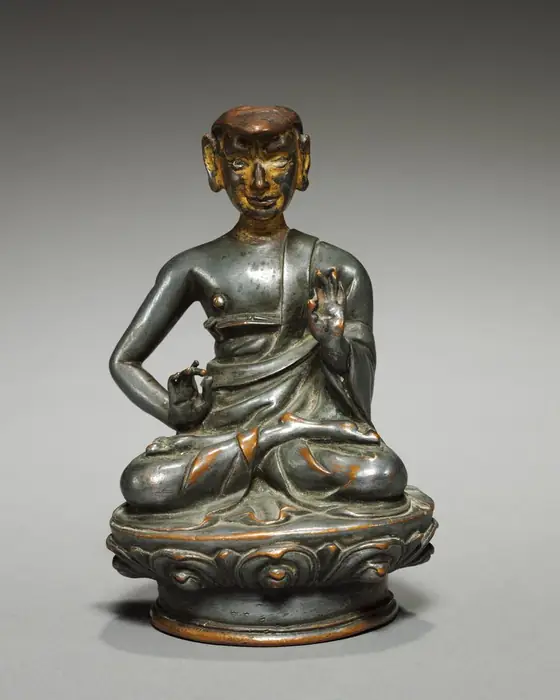 A small silver-coloured seated figure of a teacher on a lotus base, one hand raised.