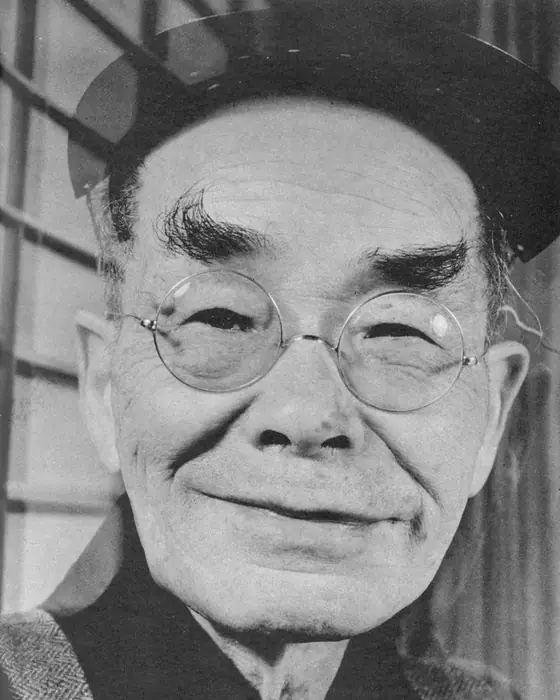 A black-and-white close portrait of a smiling old man with bushy eyebrows and round glasses.