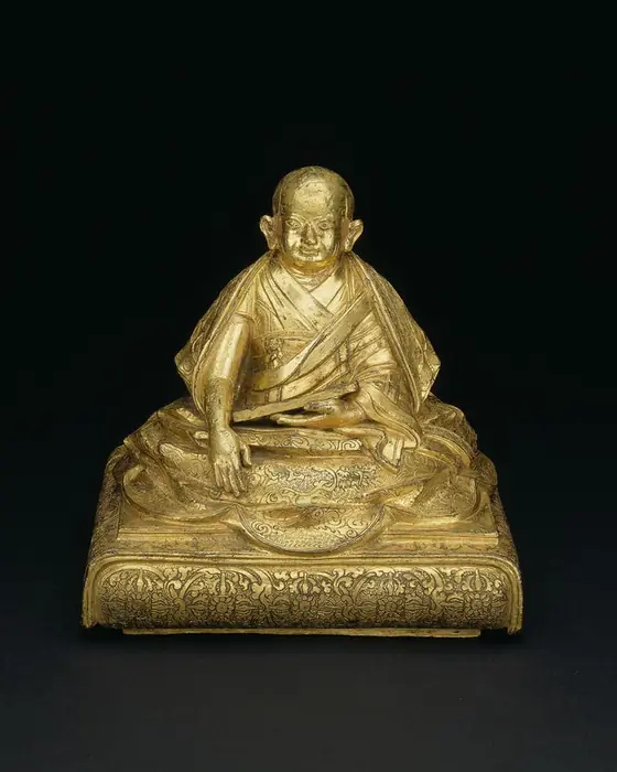 A gilded seated figure of a round-faced monk on a cushioned throne.