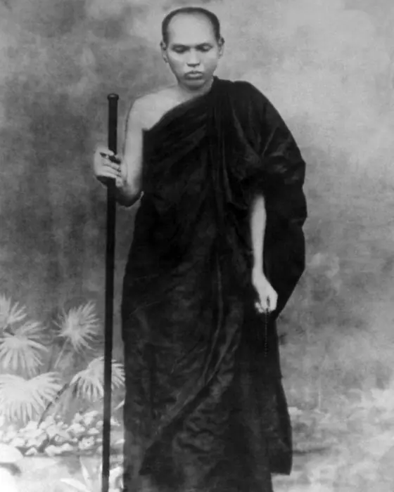A black-and-white photograph of a thin monk in a dark robe standing with a tall staff.