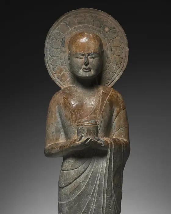 A standing stone figure of an elderly monk with a halo, holding a small cylinder before his chest.