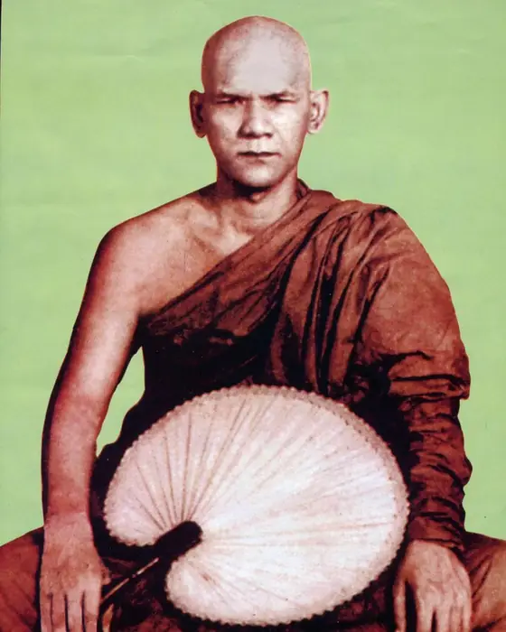 A tinted photograph of a seated monk in a maroon robe holding a large palm-leaf fan.