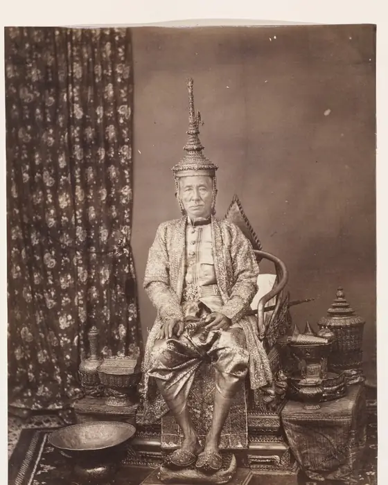 An albumen photograph of a king in a tall crown and jewelled robes, seated beside ornate vessels.