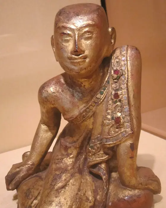 A gilded wooden figure of a kneeling monk, one hand on his knee, with red and green inlay on his robe.