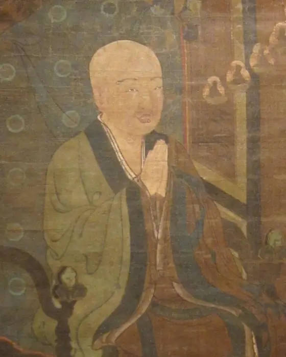 A painting of a seated monk with hands joined, in a green robe, on a chair.