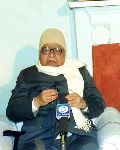A photograph of an elderly man in a white shawl and cap speaking into a microphone.