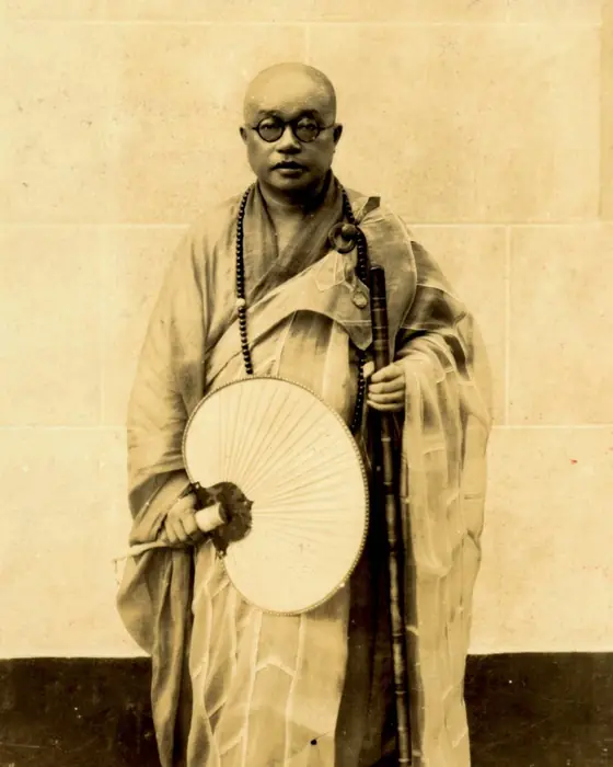 A sepia photograph of a monk in glasses and robes, holding a round fan and a staff.