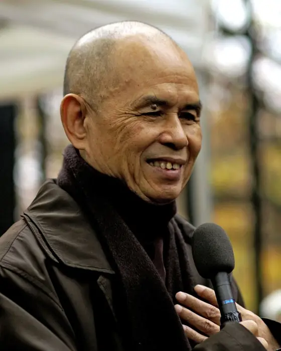 A colour photograph of a smiling elderly monk in a brown robe and dark jacket, speaking into a microphone.