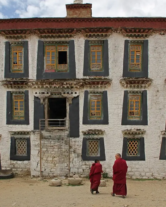 A whitewashed Tibetan monastery building with dark window frames, two monks walking in front.