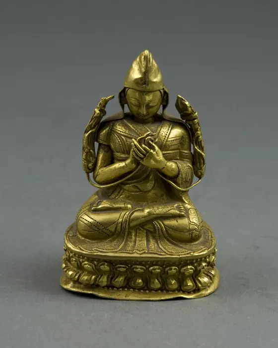 A small gold figure of a seated teacher in a pointed hat, with flowers at his shoulders.