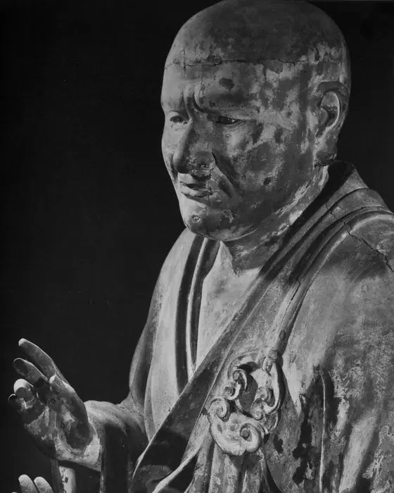 A black-and-white photograph of a wooden statue of a monk turning his head, one hand raised.