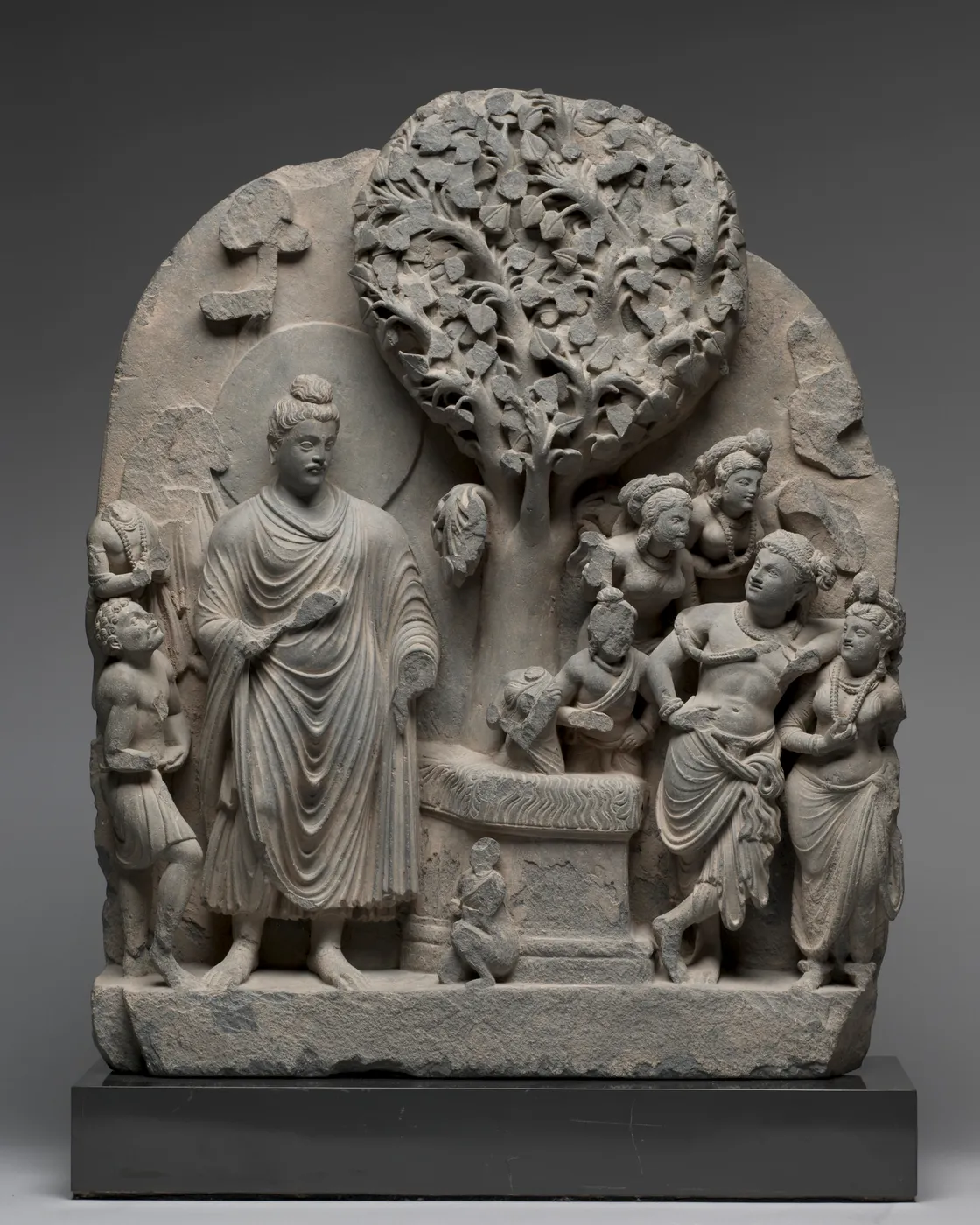 A grey stone relief with a large leafy tree at the centre, a standing robed figure and worshippers around it.