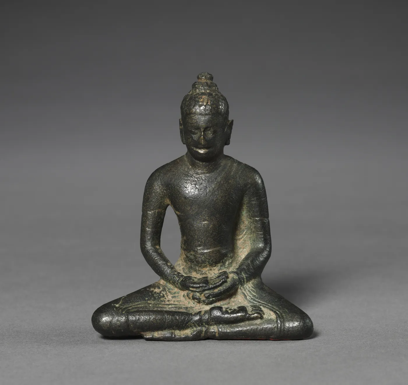 A dark bronze figure of the Buddha seated cross-legged with hands folded in his lap.
