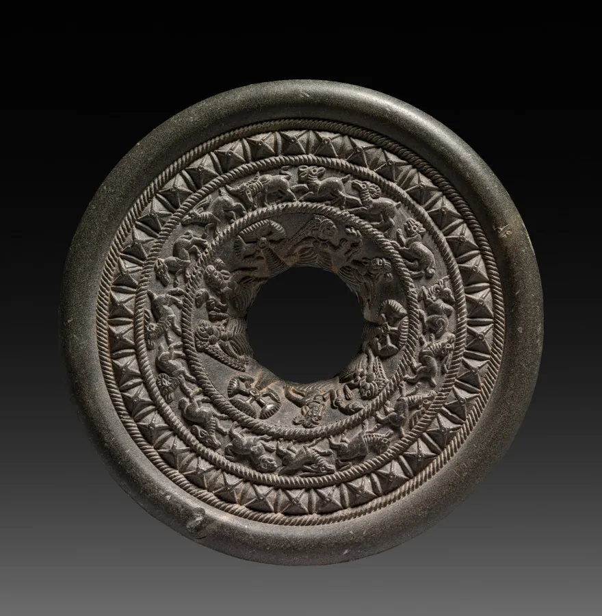 A dark stone disc with a central hole, carved in concentric bands of fine ornament.