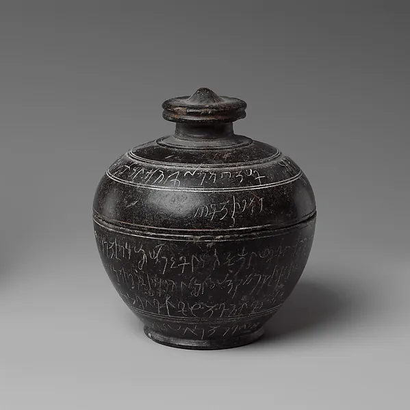 A dark rounded stone jar with a lid, its surface incised with lines of script.